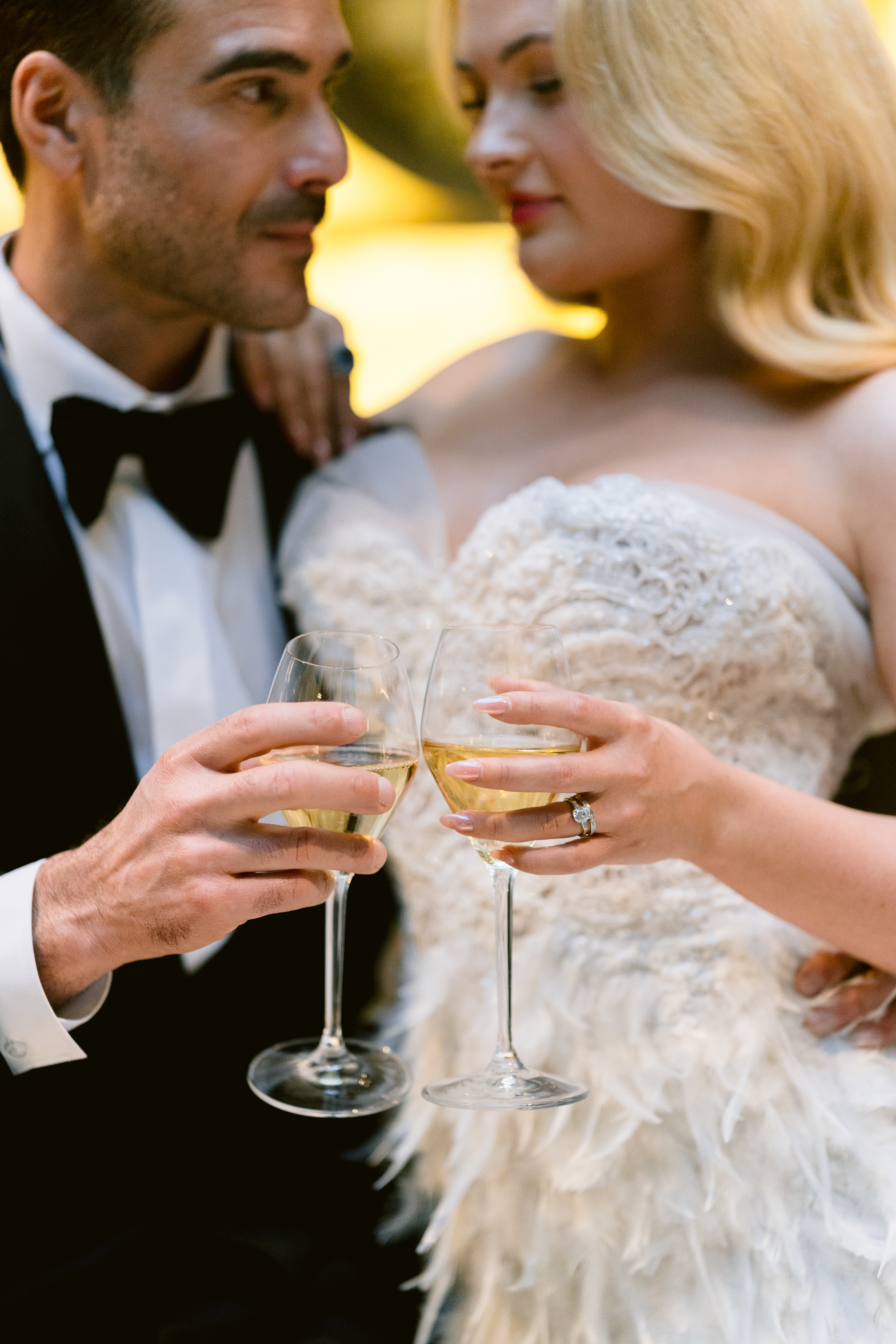 Elegant couple dressed in a tuxedo and feather-detailed bridal gown clink champagne glasses, sharing an intimate moment in warm golden light.