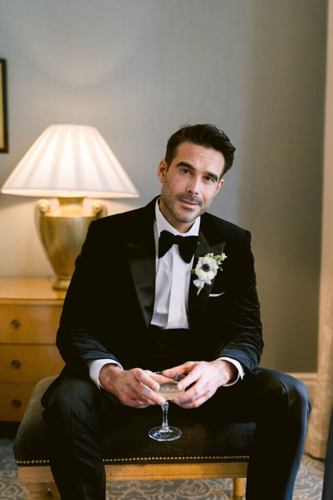 Editorial portrait of a groom holding a crystal coupe of champagne in his suite, styled for an Old Hollywood wedding at The Landmark London, featuring soft shadows, crisp tailoring, and red-carpet poise