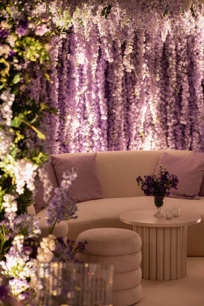 Mehndi stage with cascading lavander wisteria , ivory nabouk couch, ivory pouffe cahirs and purple floral decor on ivory coffe table