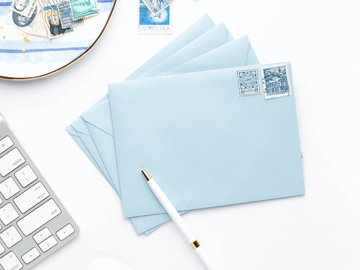 4 baby blue envelopes with white and blue stamps a whiye and gold pen on top, some more stamps in the same shade scattered on a white desk and a small white and blue plate and a silver keyboard