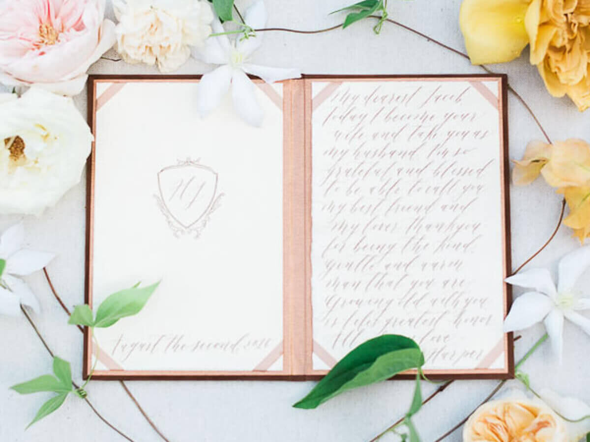 a flatlay of a fine art wedding guest book with blush decor and calligraphy, white roses and peach yellow peonies with some leaves wines