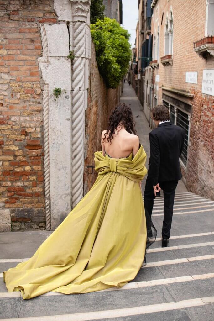 Bridesmaid in a flowing chartreuse gown walking arm in arm with the groomsman through a narrow Italian street with historic brick walls.