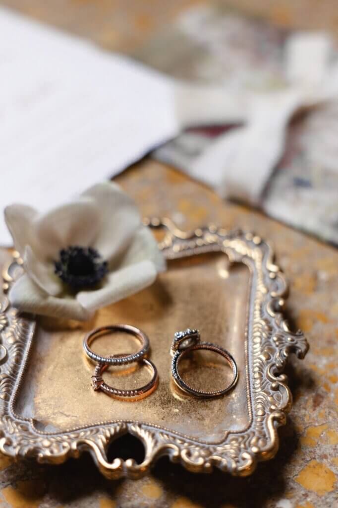 Close-up of wedding rings displayed on a vintage gold tray with a white flower, styled with soft-focus stationery in the background.