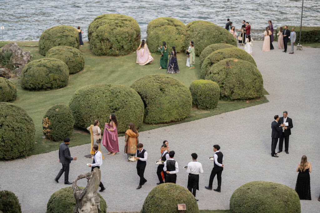 Guests mingling during a luxury lakeside wedding aperitivo in manicured Italian gardens, with uniformed staff serving drinks