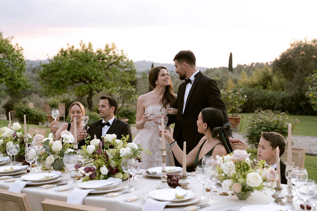 Elegant al fresco wedding dinner in Tuscany with bride and groom toasting at a long floral table, surrounded by impeccably dressed guests at golden hour

