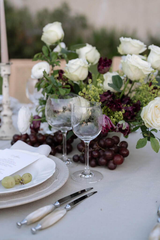 Elegant autumn wedding tablescape styled with white roses, grapes, and glassware on a neutral linen tablecloth.