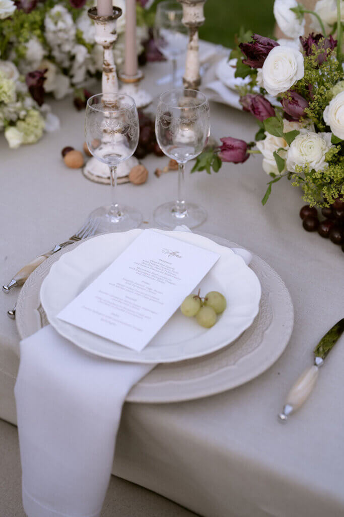 Refined autumn wedding place setting with ivory plates, menu card, and grapes styled beside crystal glassware.