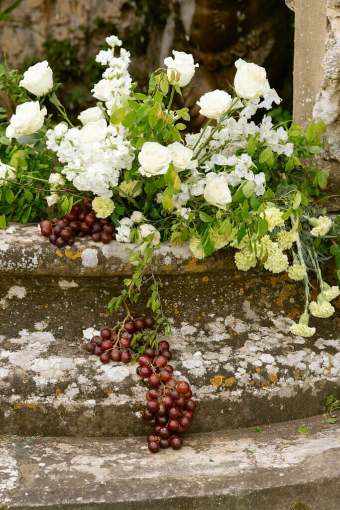 Wedding décor featuring local and seasonal florals and grapes arranged on a stone wall in Tuscany.