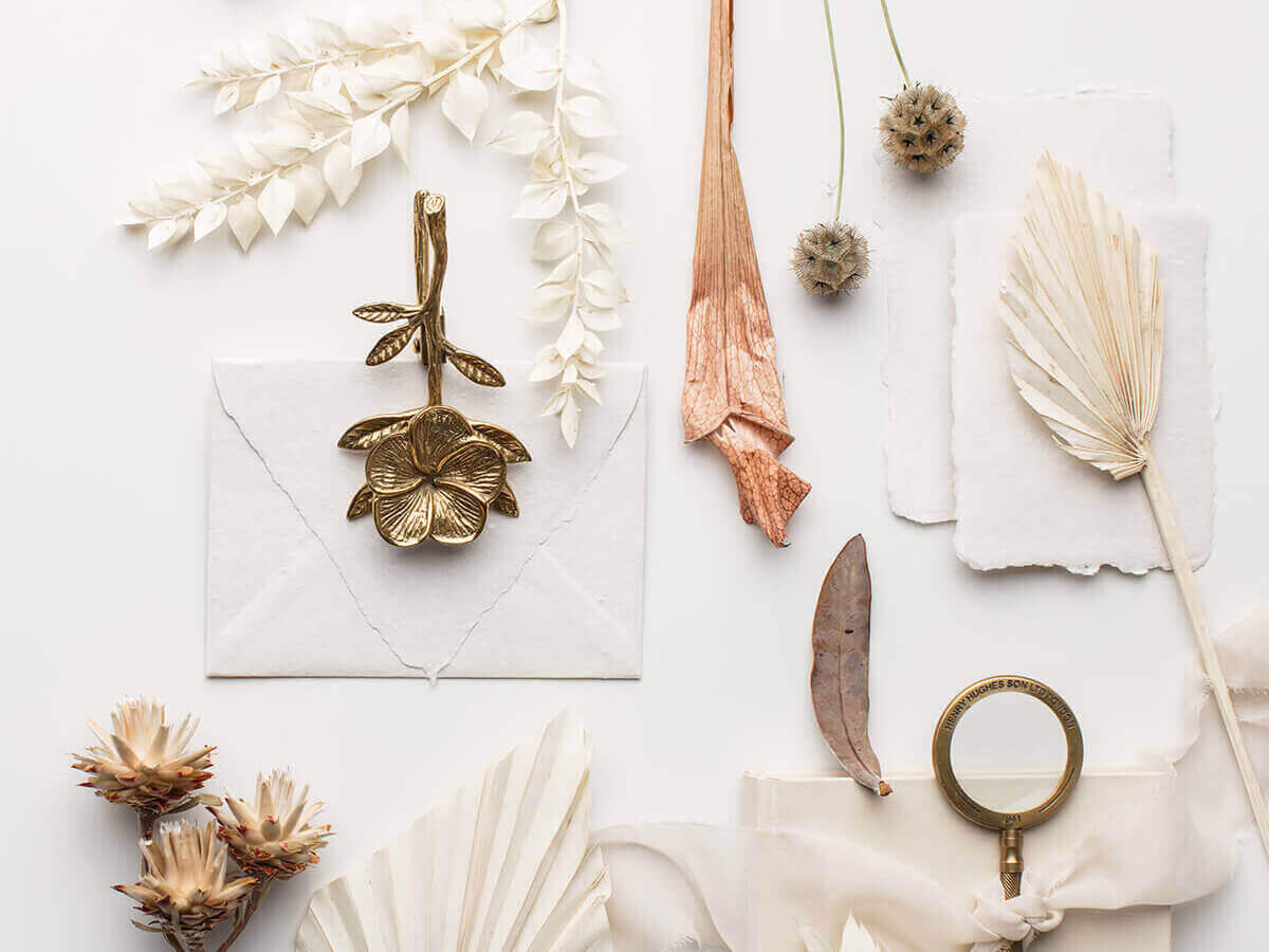 environmentally friendly wedding invitations featuring cream paper, white envelopes, some brass paper cutter and flower ornament, cream dry palm leaves, dry wild cream and rust flowers, a cream ribbon, dried rusty leaves and some cream bunny tails 